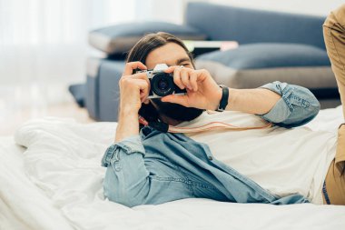 Konsantre fotoğrafçı elinde kamerayla yatakta uzanıyor ve evde fotoğraf çekiyor.