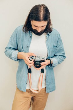 Beyaz duvarın karşısında durup elindeki kameraya bakan sıradan giysili çekici genç adam.