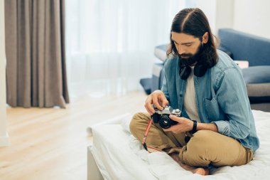 Gündelik giysiler içinde genç bir adam bir film kamerasıyla yatakta oturuyor ve ona bakıyor.