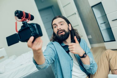 Sakallı, uzun saçlı adam, kameraya süper jest yaparken evde oturup baş parmağını kaldırıyor.