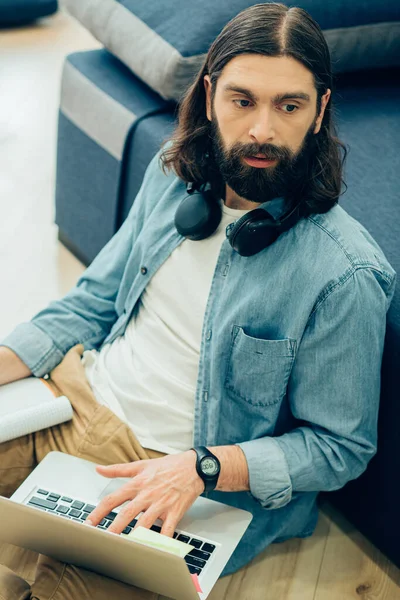 Young bearded man sitting on the floor with his back to the sofa and looking away while holding a laptop on his leg