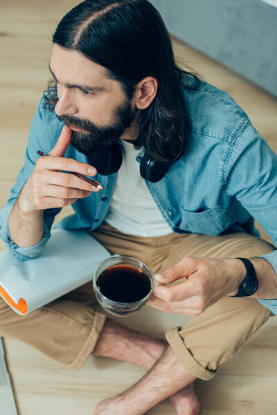 Серьезный творческий человек, сидящий на полу с чашкой кофе и трогающий губы, глядя в сторону
