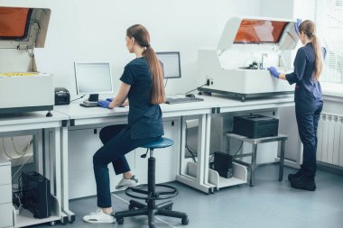 Laboratuvar bilim adamı, konsantre olmuş meslektaşı masada oturup bilgisayar kullanırken modern bir biyokimyasal analiz cihazı açıyor.