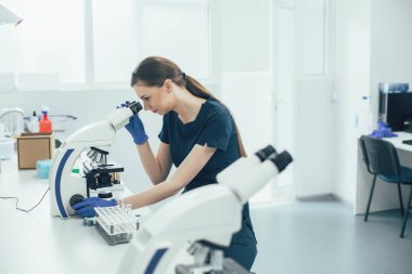 Lastik eldiven takan ve modern bir mikroskop kullanarak laboratuvarda tek başına oturan kendine güvenen bir laboratuar asistanı.