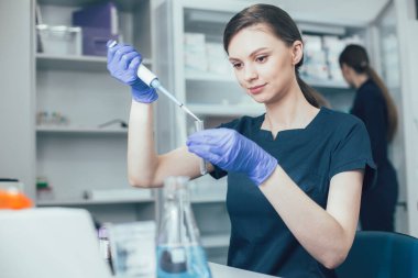 Sakin genç bir kadın laboratuarda iş yerinde oturmuş elinde bir tüp tutarak sıvıyı otomatik bir pipet kutusuyla ölçüyor.