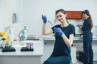 Neşeli laboratuar asistanı elinde bir test tüpüyle masada oturuyor ve manuel boru kullanırken gülümsüyor.