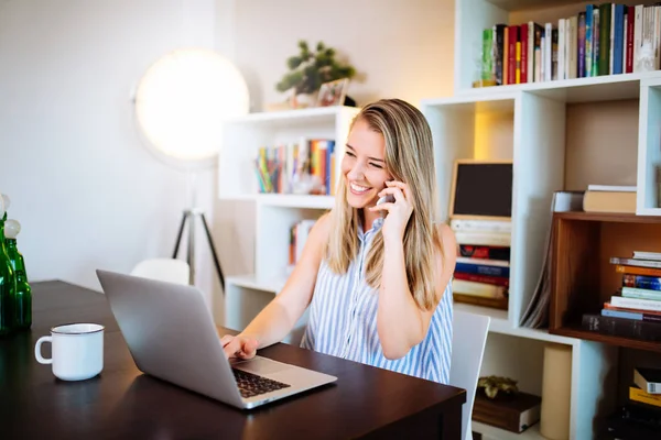 Sonriente mujer joven que trabaja en el ordenador portátil en casa mientras habla en p — Foto de Stock