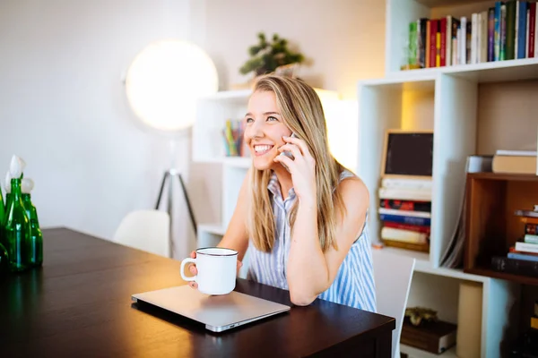 Sonriente mujer joven que trabaja en el ordenador portátil en casa mientras habla en p — Foto de Stock