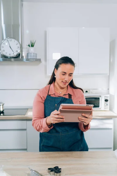 Young brunette woman checking a cookies recipe on a tablet in her kitchen for making cookies at home for the Christmas Holidays. Handmade gingerbread cookies