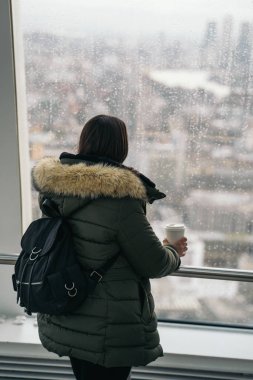 Young woman with green jacket looking at the panoramic view from the viewpoint of the London Sky Garden building. She is drinking hot coffee clipart