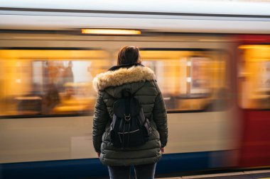 Arka planda bulanık hareket eden trenle metro istasyonunda sırt çantasından vurulan yalnız genç bir kadın.