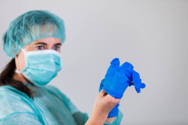 Korunaklı maskeli ve şapkalı genç bir kadın doktor koronavirüse karşı koruyucu eldiven takıyor. Kişisel koruyucu tıbbi ekipmanlar.