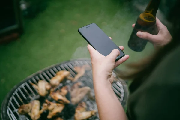 裏庭でビールを飲みながらバーベキューで鶏の羽のグリルに携帯電話で写真を撮る女性 — ストック写真