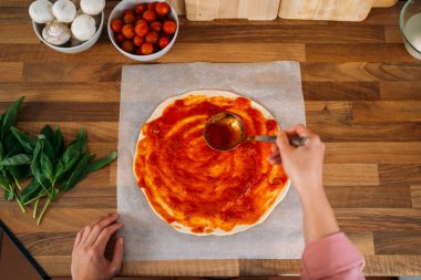 Tahta bir atölyede pizza hamuruna domates sosu ekleyen bir kadının el manzarası. Domates sosu, mozzarella, domates, fesleğen, zeytinyağı, peynir, kırsal ahşap masada servis edilen baharatlar. İtalyan pizzası.