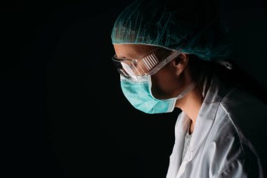 Tıbbi maske, gözlük, şapka ve virüs koruyucu giysiler takan siyah arka planda fotokopi çeken bir kadın doktoru kapatın. COVID-19 Koronavirüs salgını.