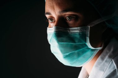 Tıbbi maske takan genç bir kadın doktor, siyah arka planda koruyucu giysiler giyip fotokopi çekiyor. COVID-19 Koronavirüs salgını.