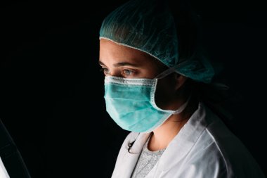 Tıbbi maske takan genç bir kadın doktor, siyah arka planda koruyucu giysiler giyip fotokopi çekiyor. COVID-19 Koronavirüs salgını.