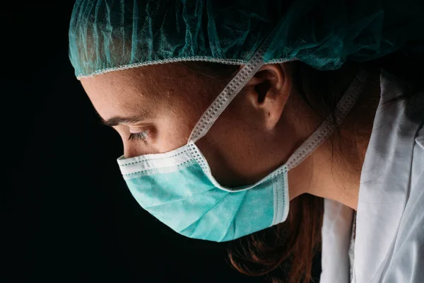 Side young woman doctor wearing medical surgical mask, cap and virus protective clothing on black background with copy space. COVID-19 The coronavirus pandemic.