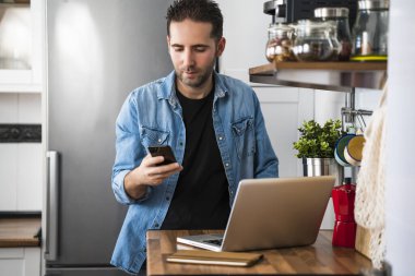 Genç adam akıllı telefonunu kullanıyor. Dairesinde dizüstü bilgisayarla çalışıyor. Evde çalışmak ve evde kalmak..