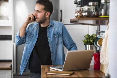 Akıllı telefonuyla konuşan ve ev mutfağında dizüstü bilgisayar kullanan bir adam. Evde çalışmak ve evde kalmak..