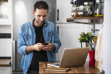Genç adam akıllı telefonunu kullanıyor. Dairesinde dizüstü bilgisayarla çalışıyor. Evde çalışmak ve evde kalmak..