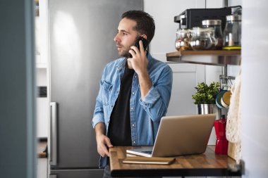 Akıllı telefonuyla konuşan ve ev mutfağında dizüstü bilgisayar kullanan bir adam. Evde çalışmak ve evde kalmak..