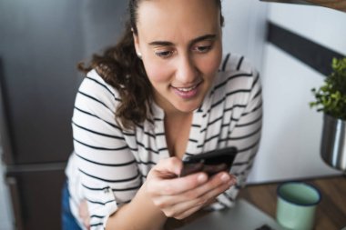 Gülümseyen genç bir kadının mutfağında akıllı telefonunu kullanırken. Evde çalışmak ve evde kalmak..