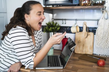 Genç bir kadın akıllı telefonunu kullanırken esniyor. Ev mutfağında dizüstü bilgisayarla çalışıyor. Evde çalış ve evde kal..