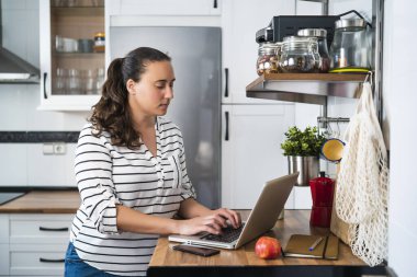 Mutfakta dizüstü bilgisayarını kullanan gülümseyen bir kadın. Evde çalışmak ve evde kalmak..