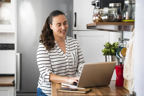 Smiling young woman working with laptop in her apartment kitchen. Working at home and stay at home.