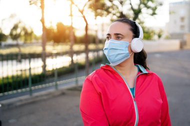 Gün batımında tıbbi maske ve kulaklık takan genç bir bayan koşucu. Covid-19 koronavirüs salgını. Corona Salgını 'nda karantinada spor ve aktif yaşam.