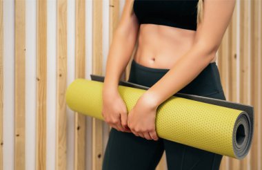 Beyaz ve ahşap duvarda yeşil yoga minderiyle duran genç ve sportif bir kadının yakın çekimi. Yoga çalışmalarından sonra dinlenme, yoga kulübünde rahat ve sosyal bir ortam..