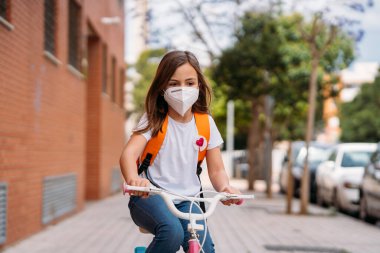 Koronavirüs salgını sırasında sokakta bisiklet süren maskeli bir kız..