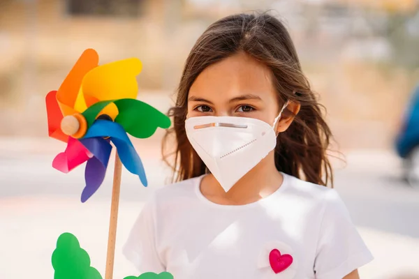 Klein meisje draagt masker en houdt een kleurrijke speelgoed windmolen, of pinwheel. — Stockfoto