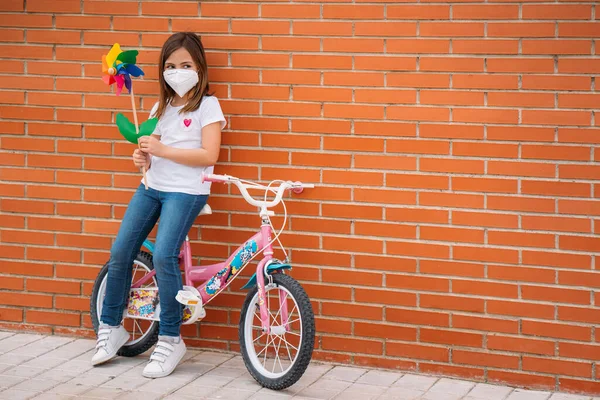 Klein meisje met een fiets dragen masker en met een kleurrijke speelgoed windmolen — Stockfoto