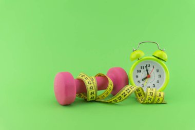 Sağlıklı yaşam tarzı konseptine başlıyorum. Yeşil arka planda pembe halterli, mezuralı ve alarmlı bir hayat.