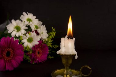 Short white candle burning on the floor in close-up among flowers against dark background. clipart