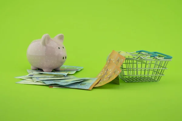 Cost of shopping or spending savings concept with banknotes scattered on a green background with a piggy bank and small wire shopping basket