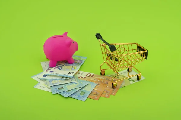Cost of shopping or spending savings concept with banknotes scattered on a green background with a piggy bank and small wire shopping cart