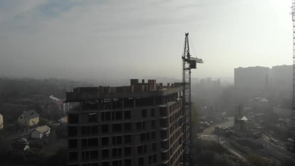 Pohled z ptačí perspektivy na věžový jeřáb v mlze stojící vedle obytné budovy. Letím nad staveništěm. Záběry dronů v reálném čase. — Stock video