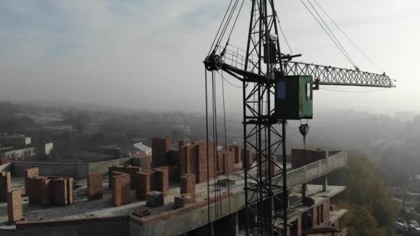 Birds eye view on tower crane in fog standing next to residential building. Flying over the construction site. Drone real time footage. — 비디오