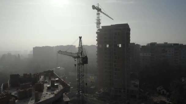 Birds eye view on tower crane in fog standing next to residential building. Flying over the construction site. Drone real time footage. — ストック動画