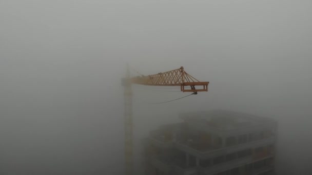 Birds eye view on tower crane boom in fog standing next to residential building. Flying over the construction site. Drone real time footage. — Stok video