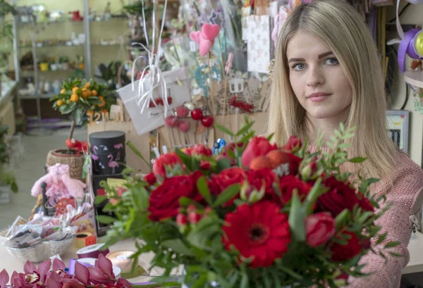 Una niña, una florista-diseñadora sostiene un hermoso ramo festivo de flores rojas y un corazón con el amor de la inscripción, especialmente hecho para el día de San Valentín, en el fondo de una floristería — Foto de Stock