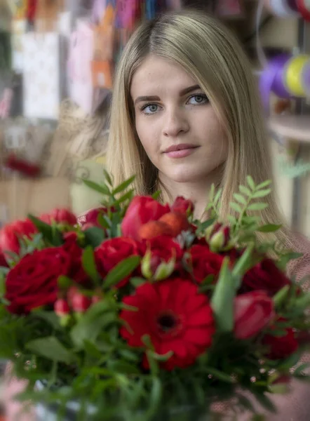 Una niña, una florista-diseñadora sostiene un hermoso ramo festivo de flores rojas y un corazón con el amor de la inscripción, especialmente hecho para el día de San Valentín, en el fondo de una floristería — Foto de Stock