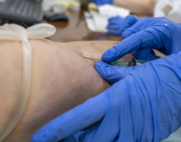 blue-gloved hands of a medical professional insert a needle into a vein to draw blood from the vein for a coronavirus test