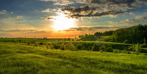 Magnífico paisaje, puesta de sol sobre campos, barrancos y bosques, cielo naranja turquesa y hierba verde brillante y hojas de árboles — Foto de Stock