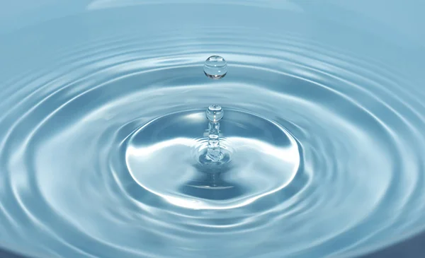 Uma gota de água cai com um respingo na água clara e azul formando círculos na água — Fotografia de Stock