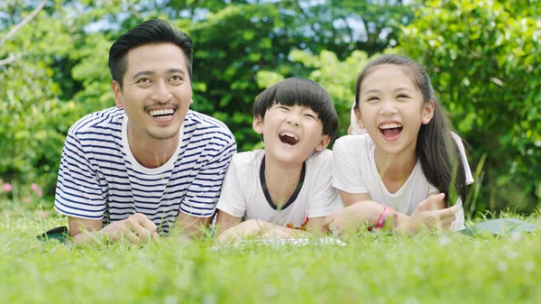 Asian family looking up & laughing when lying on stomach and reading on grassland