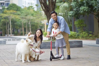 Asian parents & daughter playing scooter while walking dog in garden clipart